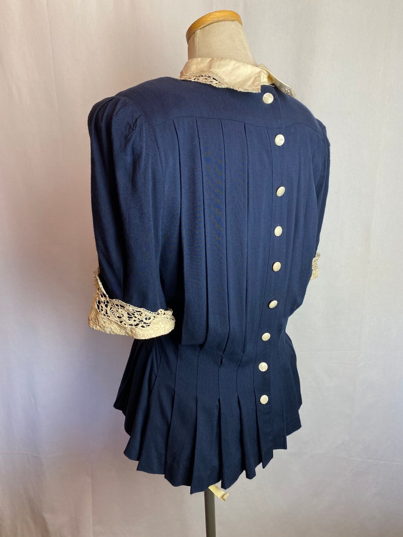 May include: A navy blue blouse with a white lace collar and cuffs. The blouse has a fitted bodice and a flared skirt. It features a row of white buttons down the back.