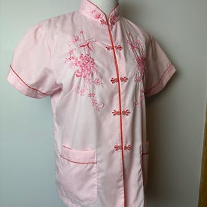 May include: A pink short-sleeved button-up shirt with red trim and floral embroidery. The shirt has a mandarin collar and a front pocket.