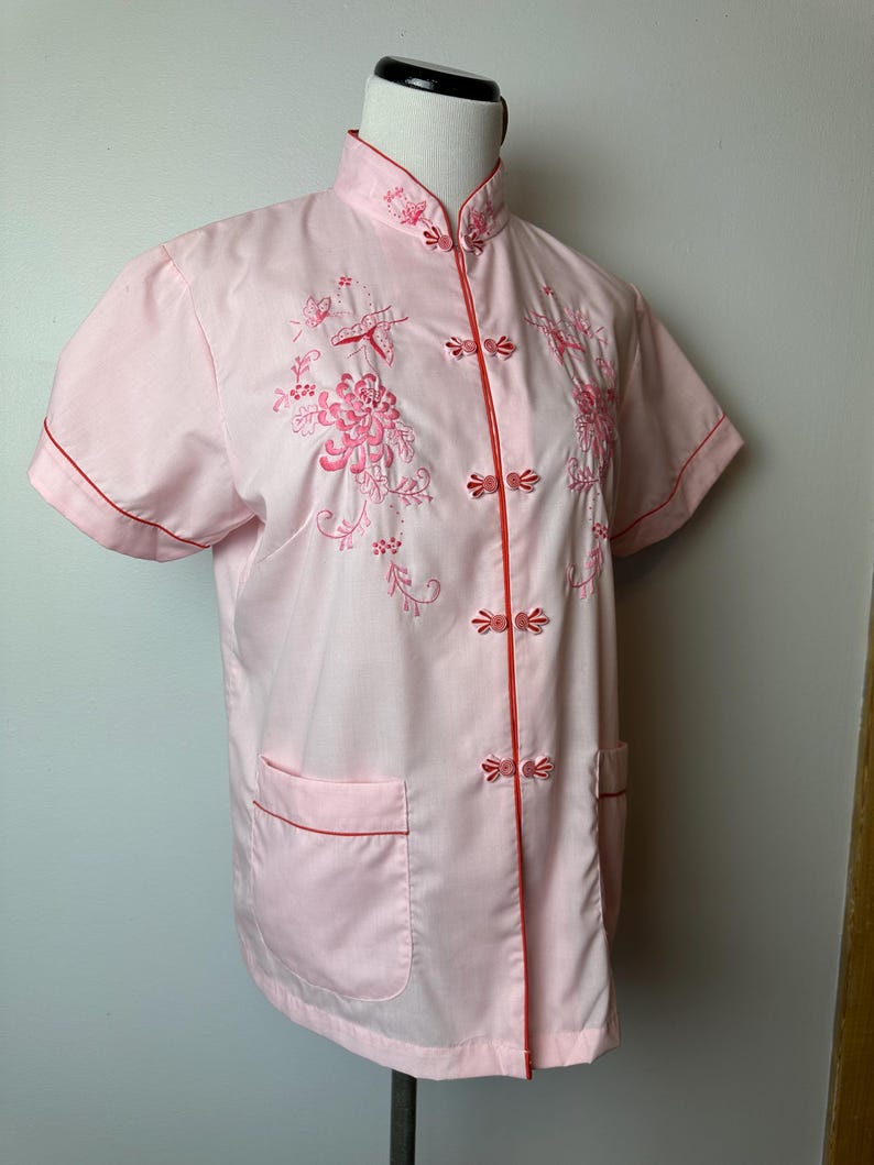 May include: A pink short-sleeved button-up shirt with red trim and floral embroidery. The shirt has a mandarin collar and a front pocket.