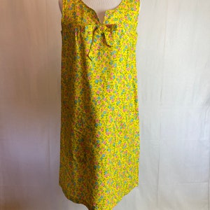 May include: A sleeveless yellow dress with a floral print and a bow at the neckline. The dress has a simple, A-line silhouette.