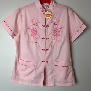 May include: A pink short-sleeved button-up shirt with red trim and floral embroidery. The shirt has a mandarin collar and two pockets. The shirt has a tag that says "Made in China".