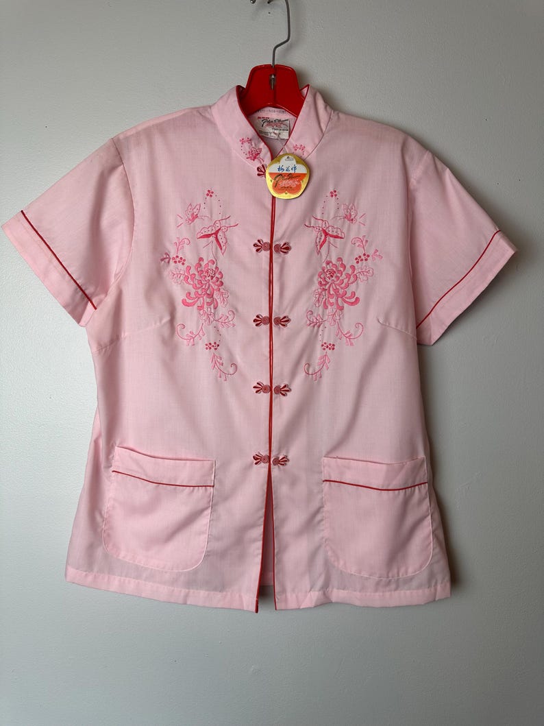 May include: A pink short-sleeved button-up shirt with red trim and floral embroidery. The shirt has a mandarin collar and two pockets. The shirt has a tag that says "Made in China".