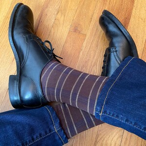 May include: A pair of black leather shoes with brown and beige striped socks. The shoes are worn and the socks are pulled up to the calves. The person is wearing blue jeans.