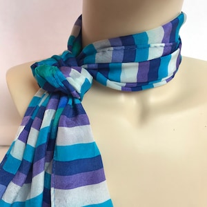 Puede incluir: Una bufanda de seda a rayas azules y moradas atada alrededor del cuello de un maniquí. La bufanda tiene un patrón de rayas azul claro, azul oscuro, morado y blanco.