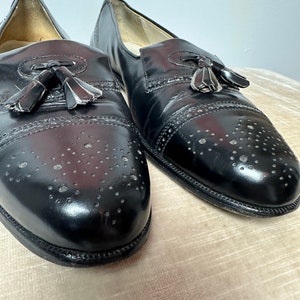 May include: A pair of black leather tassel loafers with a brogue design and perforated detailing. The shoes have a rounded toe and a slip-on style.