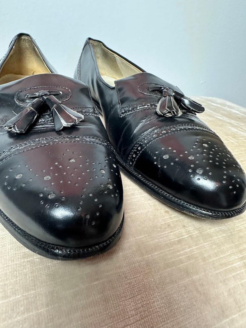 May include: A pair of black leather tassel loafers with a brogue design and perforated detailing. The shoes have a rounded toe and a slip-on style.