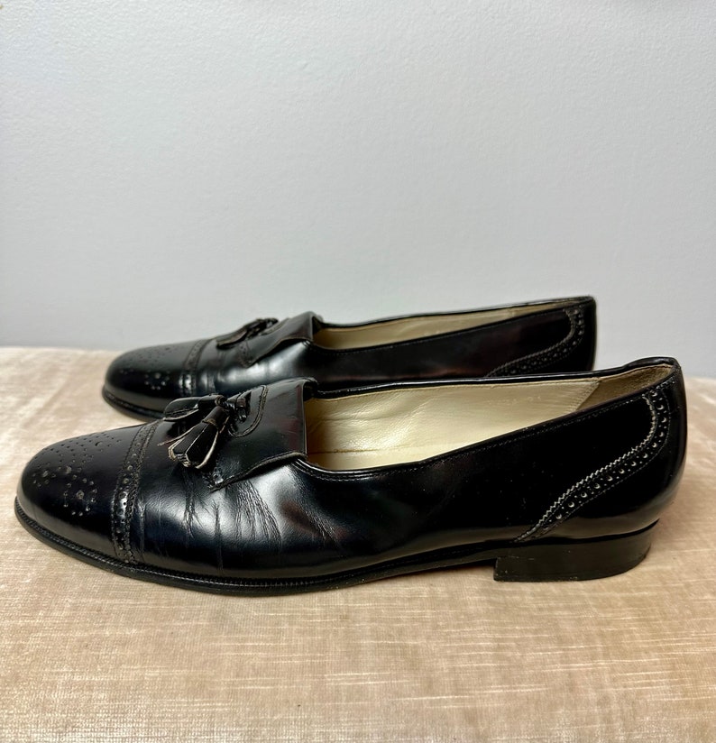 May include: A pair of black leather tassel loafers with a brogue design. The shoes have a pointed toe and a low heel.