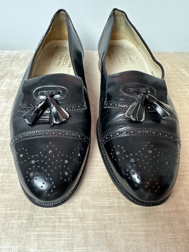May include: A pair of black leather tassel loafers with a brogue design and a perforated pattern. The shoes have a rounded toe and a slip-on style.