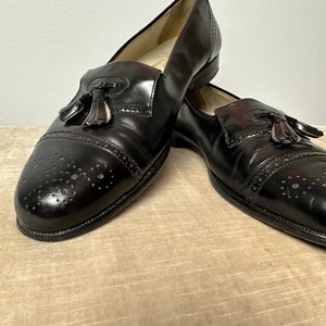 May include: A pair of black leather tassel loafers with a brogue design. The shoes have a pointed toe and a low heel.