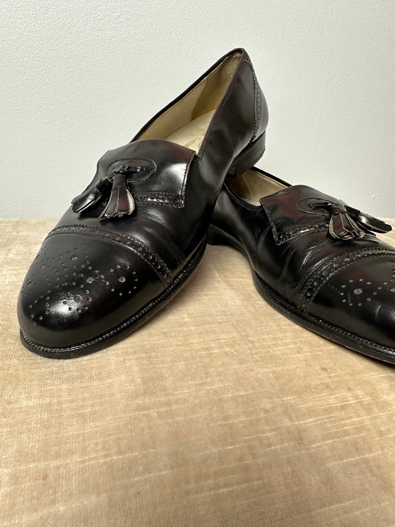 May include: A pair of black leather tassel loafers with a brogue design. The shoes have a pointed toe and a low heel.