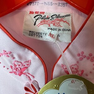 May include: A pink button-down shirt with a mandarin collar and embroidered butterflies. The shirt has a red trim and a tag that reads "Plum Blossoms" and "梅花牌".
