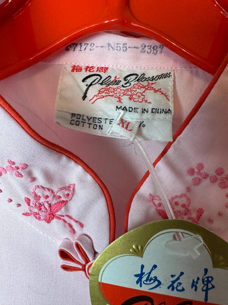 May include: A pink button-down shirt with a mandarin collar and embroidered butterflies. The shirt has a red trim and a tag that reads "Plum Blossoms" and "梅花牌".
