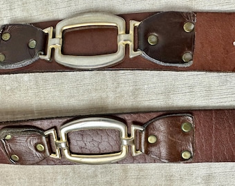 May include: Two brown leather belts with silver and gold-toned rectangular buckles. The belts have dark brown leather accents near the buckle and are secured with gold-toned rivets. The belts are displayed on a light-colored textured surface.
