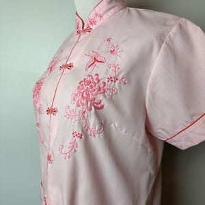 May include: A pink short-sleeved shirt with a mandarin collar and red trim. The shirt features intricate embroidery of flowers and butterflies in pink thread.