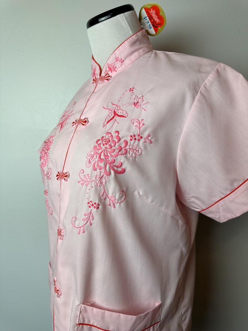 May include: A pink short-sleeved shirt with a mandarin collar and red trim. The shirt features intricate embroidery of flowers and butterflies in pink thread.