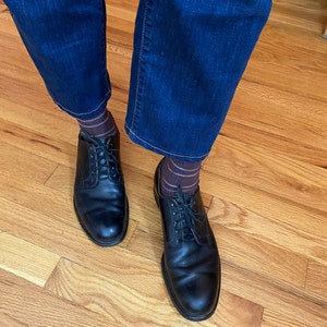 May include: A pair of black leather lace-up shoes with brown and gold striped socks. The shoes are worn by a person wearing dark blue jeans.
