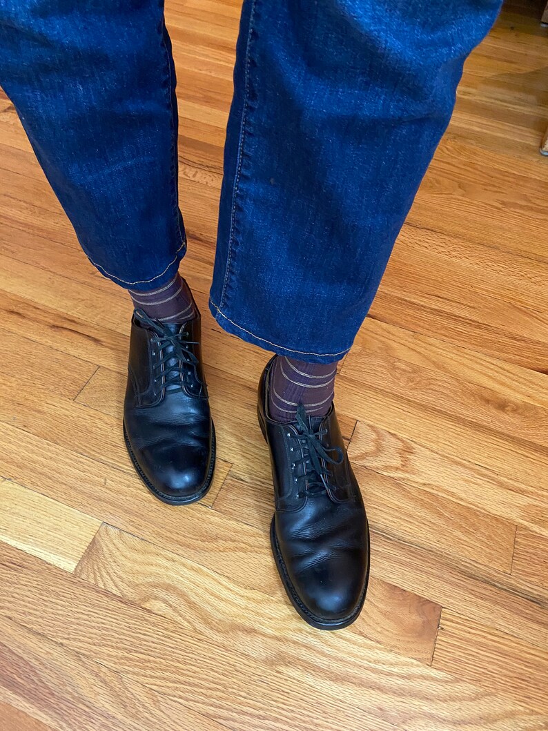 May include: A pair of black leather lace-up shoes with brown and gold striped socks. The shoes are worn by a person wearing dark blue jeans.