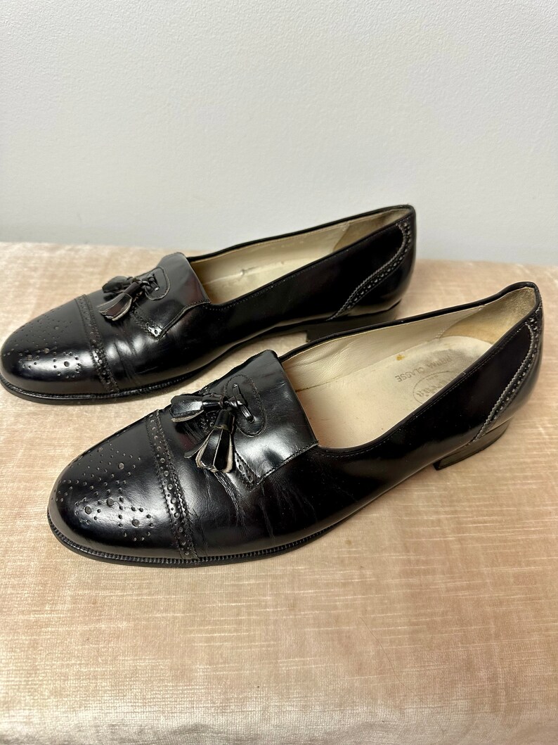 May include: A pair of black leather tassel loafers with a brogue design. The shoes have a pointed toe and a low heel.