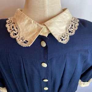 May include: A navy blue button-up shirt with a white lace trim collar and white buttons. The shirt has a gathered waist and short sleeves.