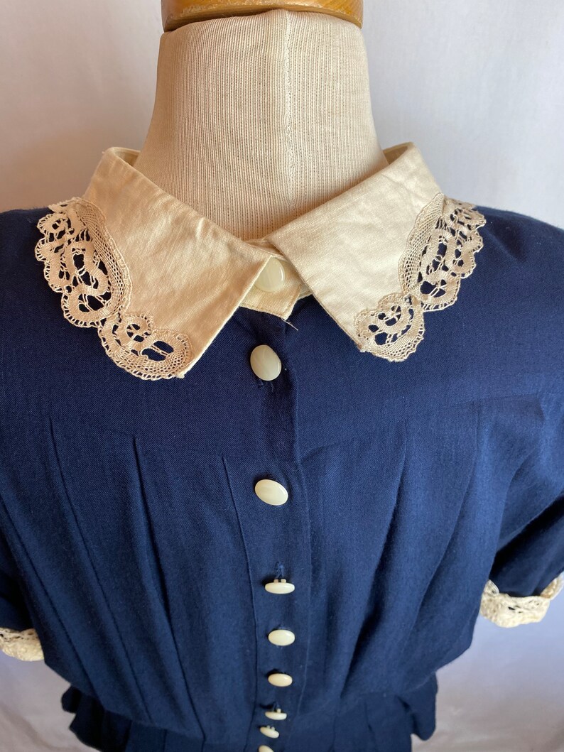 May include: A navy blue button-up shirt with a white lace trim collar and white buttons. The shirt has a gathered waist and short sleeves.
