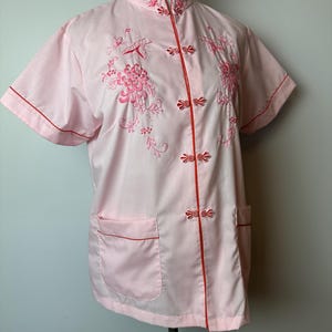 May include: A pink short-sleeved button-up shirt with red trim and floral embroidery. The shirt has a mandarin collar and a front pocket.