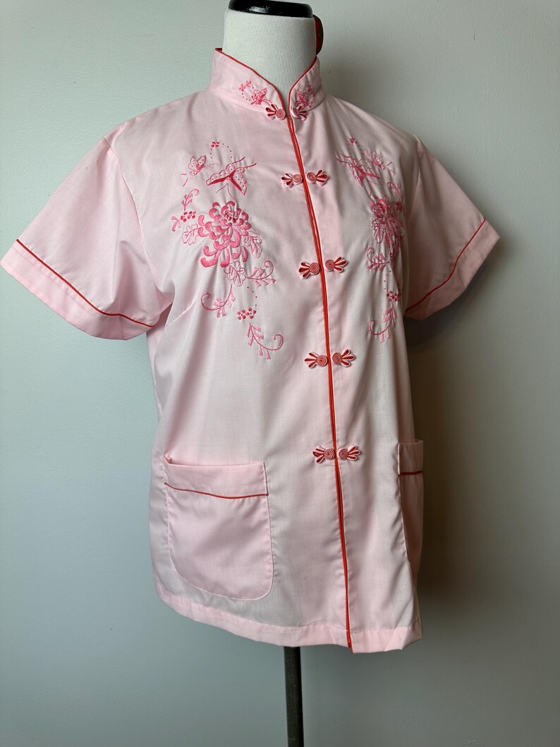 May include: A pink short-sleeved button-up shirt with red trim and floral embroidery. The shirt has a mandarin collar and a front pocket.