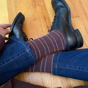 May include: A pair of black leather shoes with brown and white striped socks and blue jeans. The shoes have laces and are worn on a wooden floor.