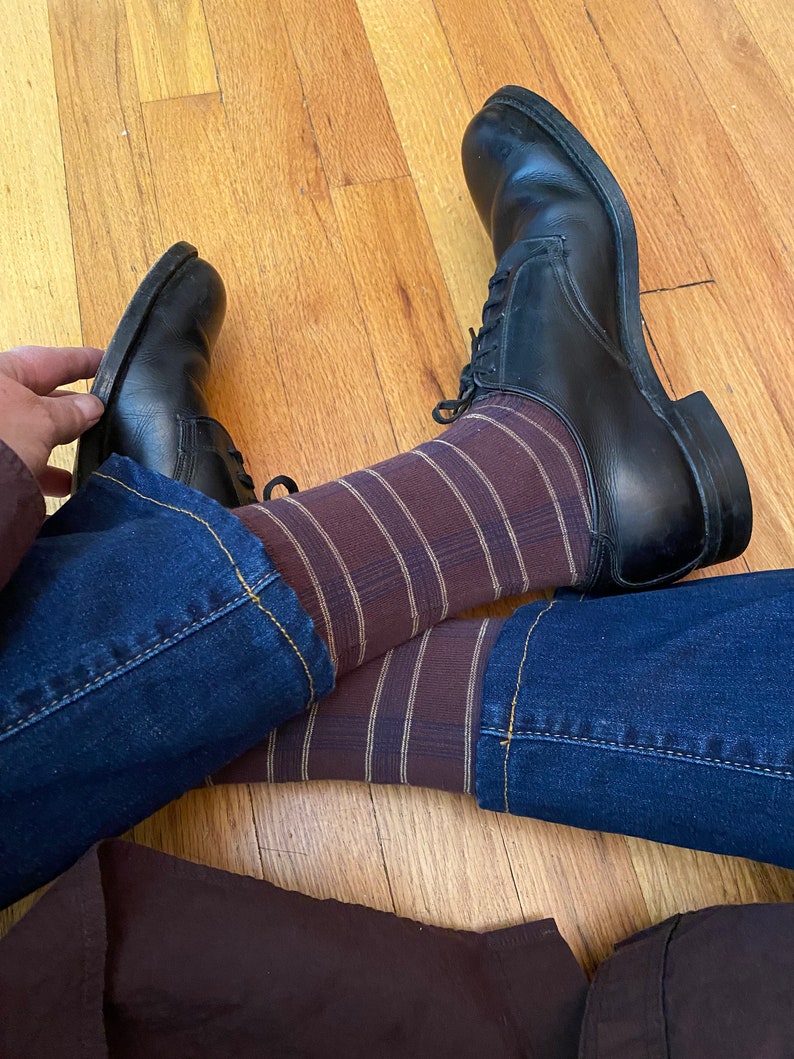 May include: A pair of black leather shoes with brown and white striped socks and blue jeans. The shoes have laces and are worn on a wooden floor.