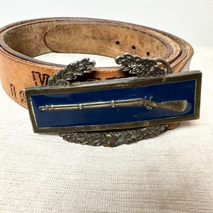 Puede incluir: Un cinturón de cuero marrón con una hebilla rectangular. La hebilla tiene un fondo azul y un diseño de rifle dorado, enmarcado con metal plateado y elementos decorativos. El cinturón está usado y tiene algo de texto.