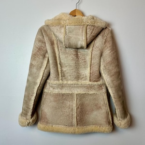 May include: A tan faux suede coat with a hood, displayed from the back. The coat features a light beige faux fur lining on the hood, cuffs, and interior. The coat has a belt detail at the waist and is hanging on a wooden hanger.