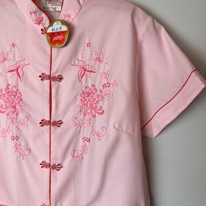 May include: A pink short-sleeved shirt with a mandarin collar and red trim. The shirt features embroidered floral and butterfly designs. The shirt has a tag that reads "Made in Hong Kong".