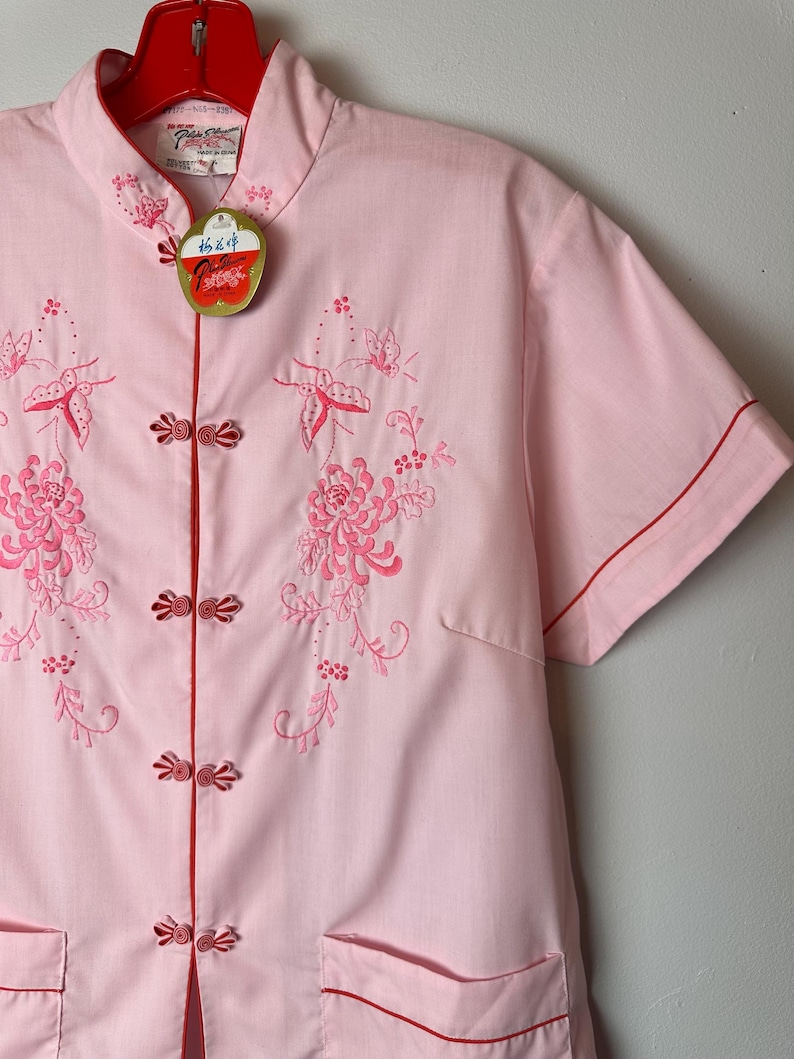 May include: A pink short-sleeved shirt with a mandarin collar and red trim. The shirt features embroidered floral and butterfly designs. The shirt has a tag that reads "Made in Hong Kong".