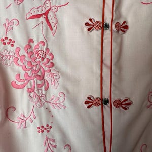 May include: A white button-up shirt with pink floral embroidery and red fabric buttons. The shirt has a mandarin collar and a red trim along the front.