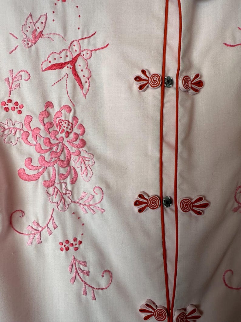 May include: A white button-up shirt with pink floral embroidery and red fabric buttons. The shirt has a mandarin collar and a red trim along the front.