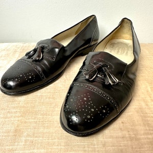 May include: A pair of black leather tassel loafers with brogue detailing. The shoes have a pointed toe and a slip-on design.