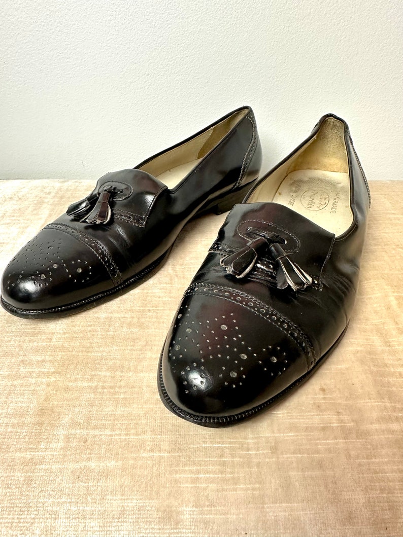 May include: A pair of black leather tassel loafers with brogue detailing. The shoes have a pointed toe and a slip-on design.
