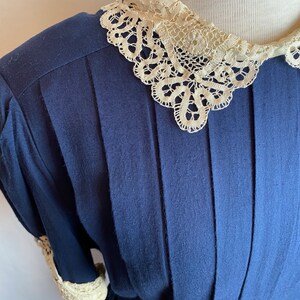 May include: A navy blue dress with a white lace collar and cuffs. The dress has a pleated bodice and a fitted waist.