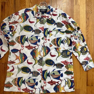 May include: A white long-sleeved button-down shirt with a colorful fish print. The shirt has a pocket on the left side.