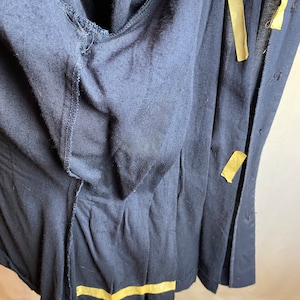 May include: A dark blue garment with a button closure and a seam detail. The garment is folded in half and has a piece of yellow tape along the bottom edge.