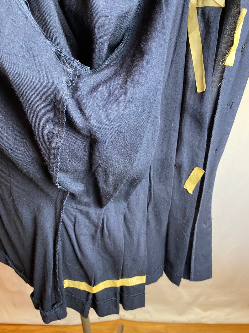 May include: A dark blue garment with a button closure and a seam detail. The garment is folded in half and has a piece of yellow tape along the bottom edge.