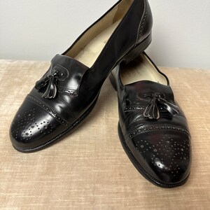 May include: A pair of black leather tassel loafers with a brogue design and a perforated pattern. The shoes have a rounded toe and a low heel.