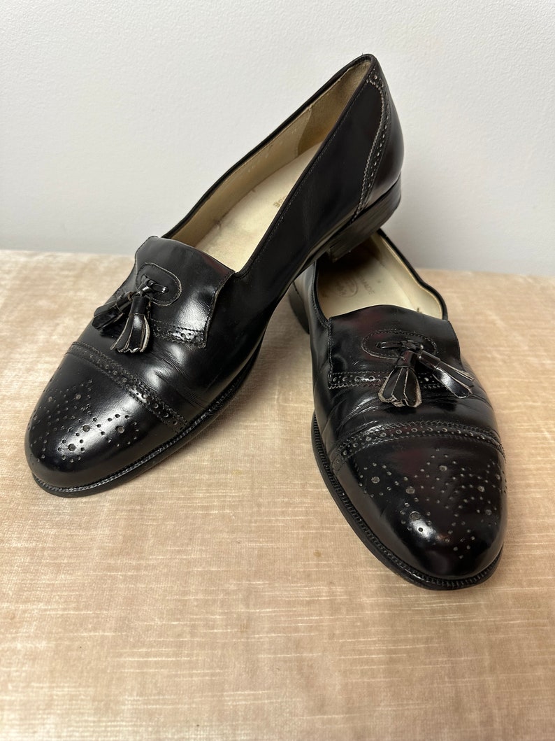 May include: A pair of black leather tassel loafers with a brogue design and a perforated pattern. The shoes have a rounded toe and a low heel.