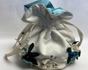 Puede incluir: Una bolsa de cordón blanco con bordado floral azul y verde. La bolsa tiene un forro de satén azul y dos correas blancas.