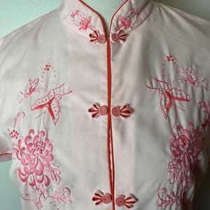 May include: A pink short-sleeved shirt with a mandarin collar and red trim. The shirt features pink floral embroidery with butterflies and a button closure.