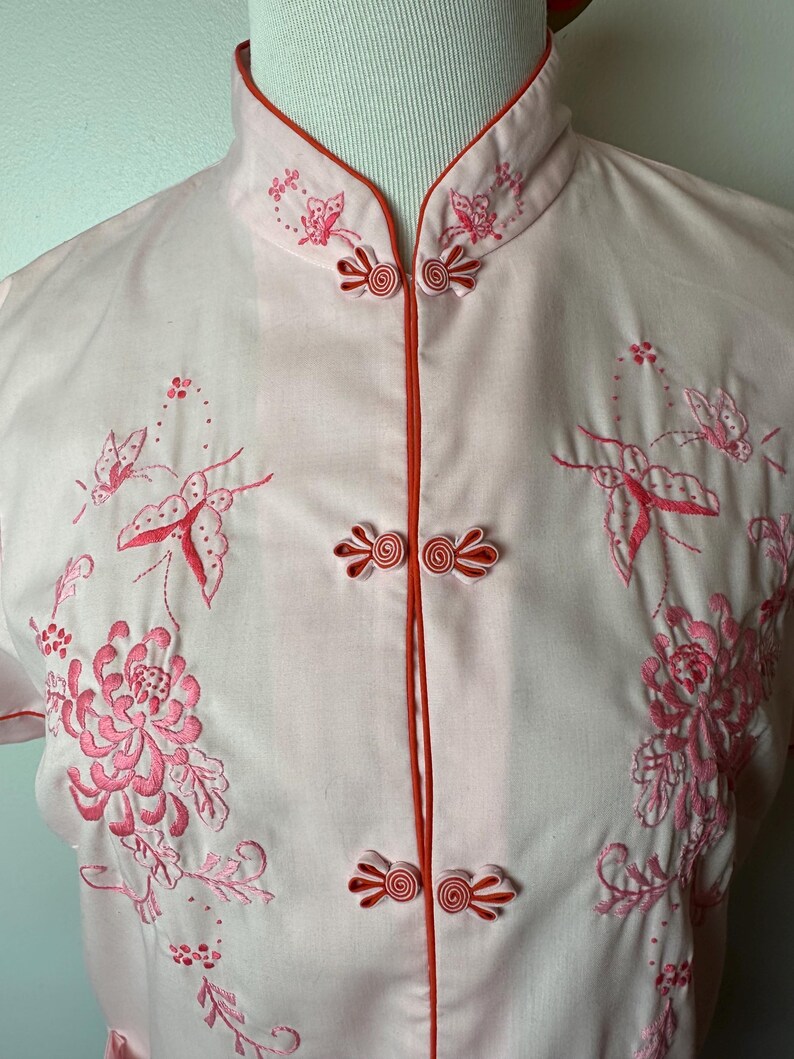 May include: A pink short-sleeved shirt with a mandarin collar and red trim. The shirt features pink floral embroidery with butterflies and a button closure.