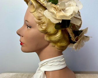 May include: A vintage straw hat with a wide brim, adorned with cream-colored fabric flowers and green leaves. The hat is displayed on a mannequin head with a white scarf. The mannequin has blonde hair and red lipstick.