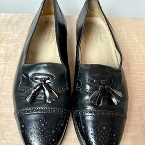 May include: A pair of black leather tassel loafers with a brogue design and perforated detailing. The shoes have a slip-on style and are made in Italy.