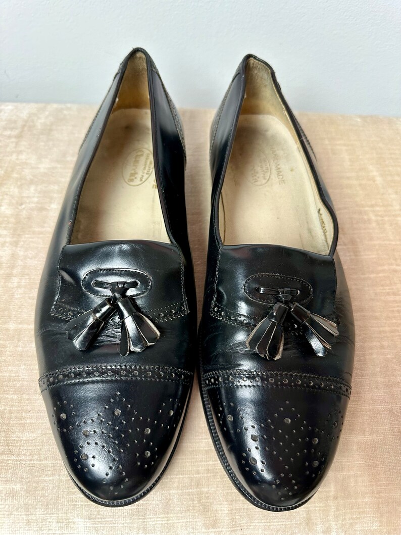 May include: A pair of black leather tassel loafers with a brogue design and perforated detailing. The shoes have a slip-on style and are made in Italy.