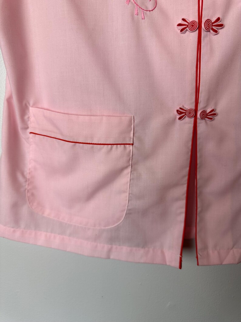 May include: A pink button-up shirt with red trim and a pocket. The shirt has a traditional Chinese-style collar and buttons.