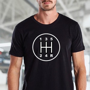May include: Black t-shirt with a white graphic of a car's gear shift pattern. The pattern includes the numbers 1, 2, 3, 4, 5, and R.